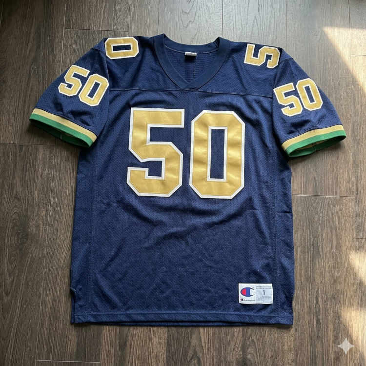 A flat-lay photograph of a navy blue mesh football jersey on a dark wood-plank floor. The jersey features the number '50' in large gold block digits with a white outline on the chest, shoulders, and sleeves. The sleeve cuffs are accented with gold and green horizontal stripes, and a Champion jock tag is visible at the bottom right.