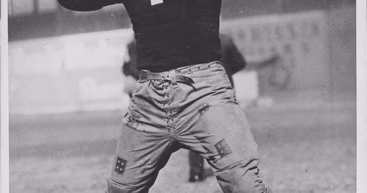 A full-length vintage photograph of Benny Friedman from 1926 during his collegiate career at the University of Michigan. He is shown in an athletic pose with one arm raised, wearing a dark long-sleeved jersey with reinforced canvas patches on the chest and shoulders. He is wearing quilted, lace-up trousers and high-top leather cleats against a plain light background.