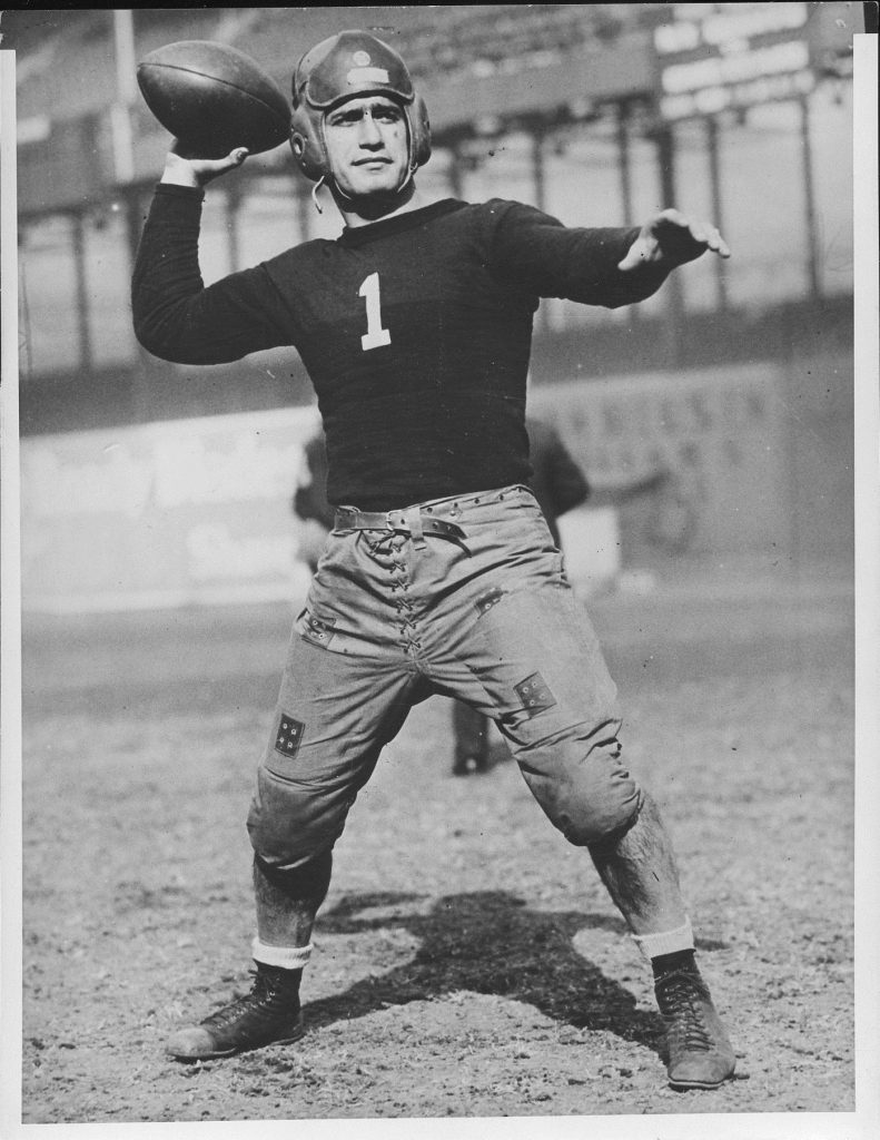 A full-length vintage photograph of Benny Friedman from 1926 during his collegiate career at the University of Michigan. He is shown in an athletic pose with one arm raised, wearing a dark long-sleeved jersey with reinforced canvas patches on the chest and shoulders. He is wearing quilted, lace-up trousers and high-top leather cleats against a plain light background.