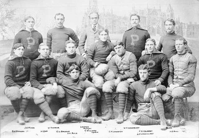 The black-and-white archival photo features 13 members of the 1895 Penn team posed in front of a gothic collegiate building. The players wear heavy wool sweaters emblazoned with a large block "P," quilted canvas trousers, and high-top leather boots. A team manager in a formal suit stands in the back row, and a player in the center holds a football marked "95". The names of the players, including Hall of Famers like George Brooke and Truxtun Hare, are printed along the bottom edge.