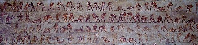 The image is a multi-tiered sequence of "action shots" featuring pairs of wrestlers in various stages of a match. The figures are painted in alternating dark red and black/brown tones to help the viewer distinguish between the two competitors in a clinch. They are shown performing recognizable maneuvers: leg trips, waist locks, lifts, and pins. The repetitive, frame-by-frame nature of the mural mimics a modern-day film strip or a "Football Archaeology" diagram of line play.