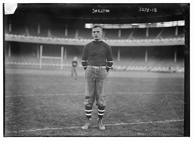 Vintage 1914 archival portrait of Cornell Big Red football player Arthur 'Art' Shelton. Black and white full-body shot showing Shelton in a dark woolen turtleneck sweater with sleeve stripes and canvas padded pants. A primary source for Bain News Service sports history and Pigskin Dispatch archives.
