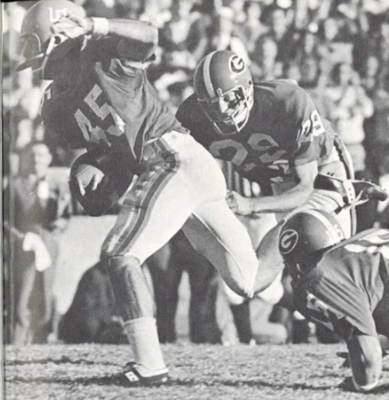 A black-and-white action photograph of Florida Gators wide receiver Carlos Alvarez (#45) from 1971. Captured mid-stride, Alvarez is shown eluding a Georgia Bulldogs defender who is diving for a tackle. Another Georgia player (#15) is visible on the ground in the foreground. The stadium background is filled with a blurred, sun-drenched crowd, and Alvarez’s classic 'UF' helmet and striped jersey details are prominent.