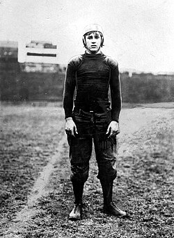 A full-length black-and-white archival photograph of a vintage football player standing on a grass field. He is wearing a long-sleeved, dark compression-style jersey with reinforced padded shoulders and dark, bulky padded trousers. He wears a classic flat-top leather helmet with ear flaps. A faint yard line is visible on the turf behind him, and the background shows a blurred, dark stadium structure under a pale sky.