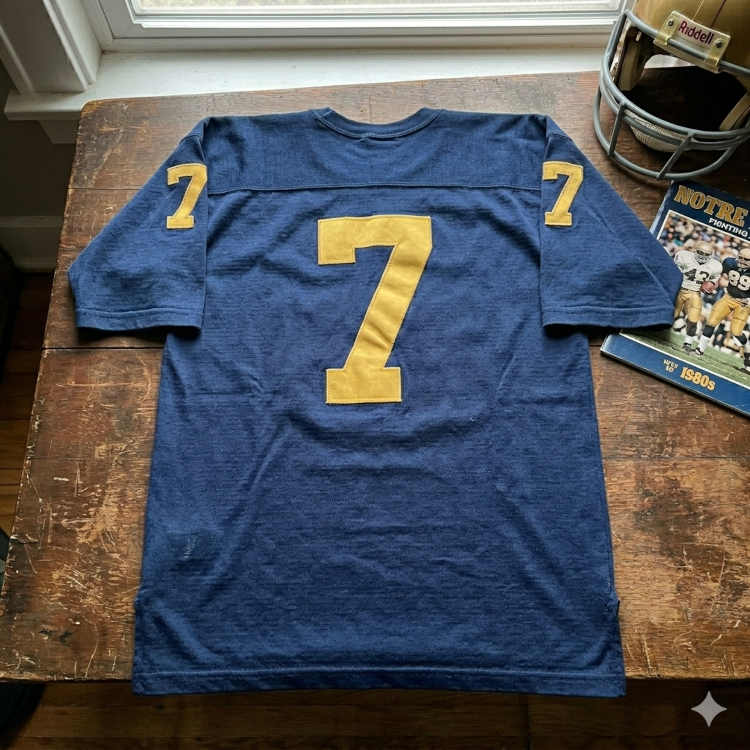 The center of the display features a navy blue, heavy-knit cotton or wool-blend jersey with the number 7 rendered in large, gold felt digits on the back and matching smaller numbers on the sleeves. The jersey is laid flat on a rustic wooden table, accompanied by a 1980s Notre Dame vs. Michigan State game program and a gold Riddell suspension helmet with a grey facemask.