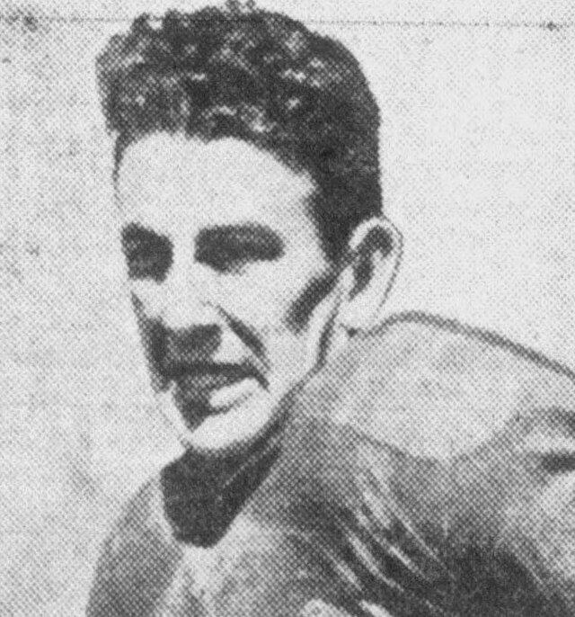 This grainy halftone portrait is a quintessential artifact of early 20th-century sports media, likely depicting a prominent player from the late 1920s or 1930s. Option 1: Descriptive & Informative (Best for general use) "A high-contrast, black-and-white halftone headshot of a vintage football player. The man has short, curly dark hair and a determined expression. He is wearing a football jersey with visible reinforced shoulder padding. The image has a heavy 'dot' texture, characteristic of newspaper or program printing from the mid-20th century.