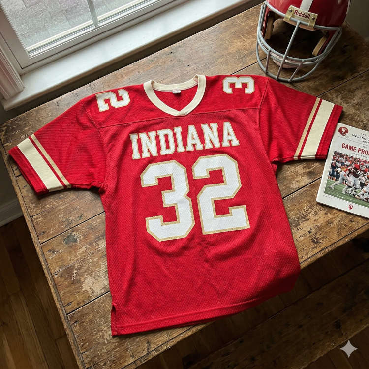 1980s-era red Indiana #32 jersey display featuring a vintage game program and a classic Hoosiers helmet on a rustic wood background.