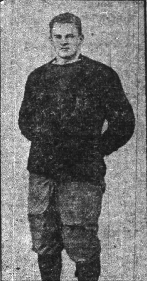 A grainy, black-and-white archival photograph of Navy Midshipmen fullback Jack Dalton, circa 1910. The full-length portrait shows Dalton standing with his hands behind his back, wearing a heavy, dark, long-sleeved wool sweater-style jersey and light-colored padded canvas trousers. His legs are encased in dark stockings. The image has the high-contrast, textured quality typical of early 20th-century newsprint or yearbook photography.