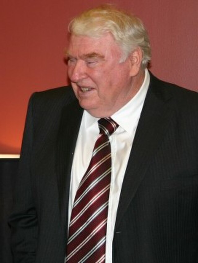 Archival photograph of NFL legend John Madden. Madden is shown in a professional suit and striped tie, smiling while looking off-camera. A primary source for historical research into coaching, broadcasting, and football cultural history for Pigskin Dispatch archives.