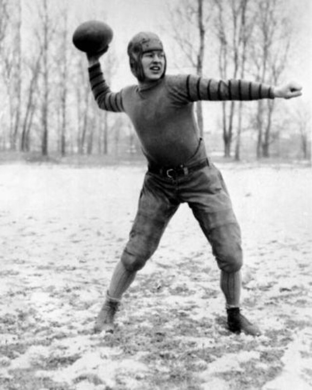 The black-and-white photograph shows a young Lambeau in a dynamic passing pose on a snowy, mud-streaked field. He is wearing a dark, form-fitting jersey with striped sleeves, characteristic of the early "Iron Man" era. His equipment includes quilted trousers, high-top leather boots, and a primitive, soft-shell leather helmet. The background of bare trees and a dusting of snow perfectly captures the rugged, winter-bound identity of football in Wisconsin.