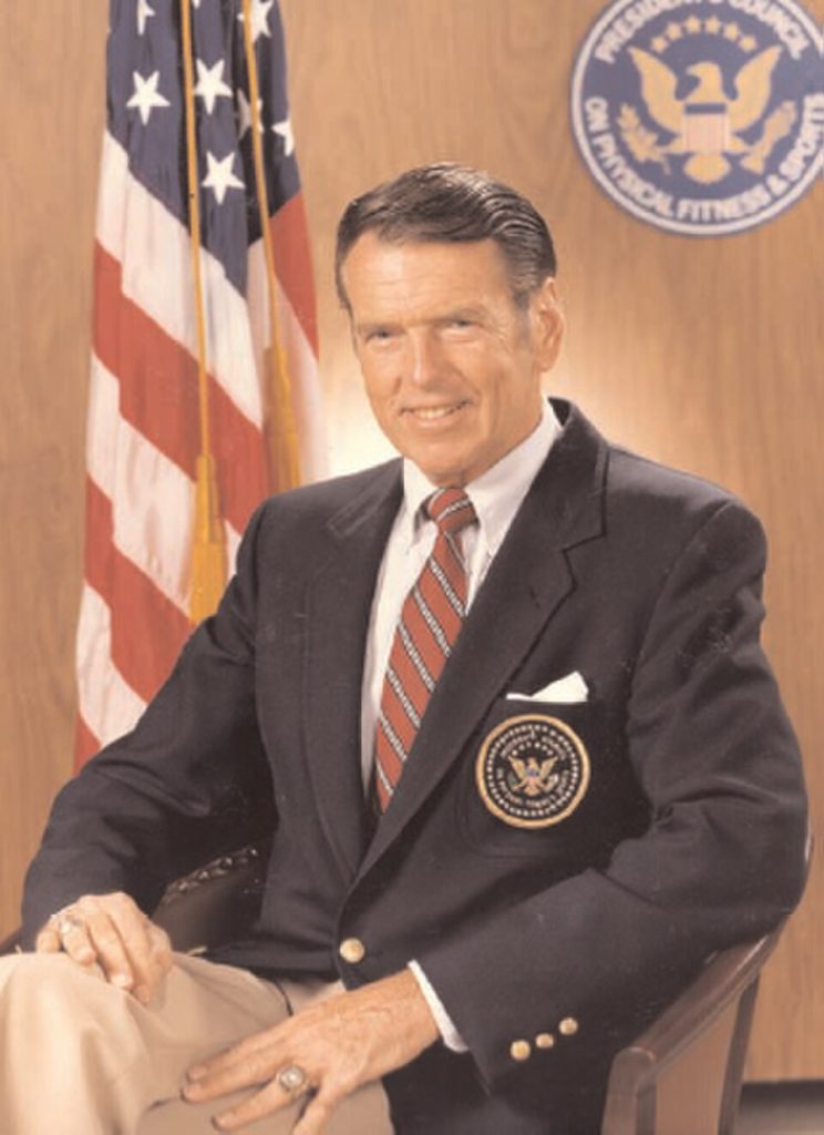 Color publicity photo of George Allen posing in a black sports coat in front of an American flag.