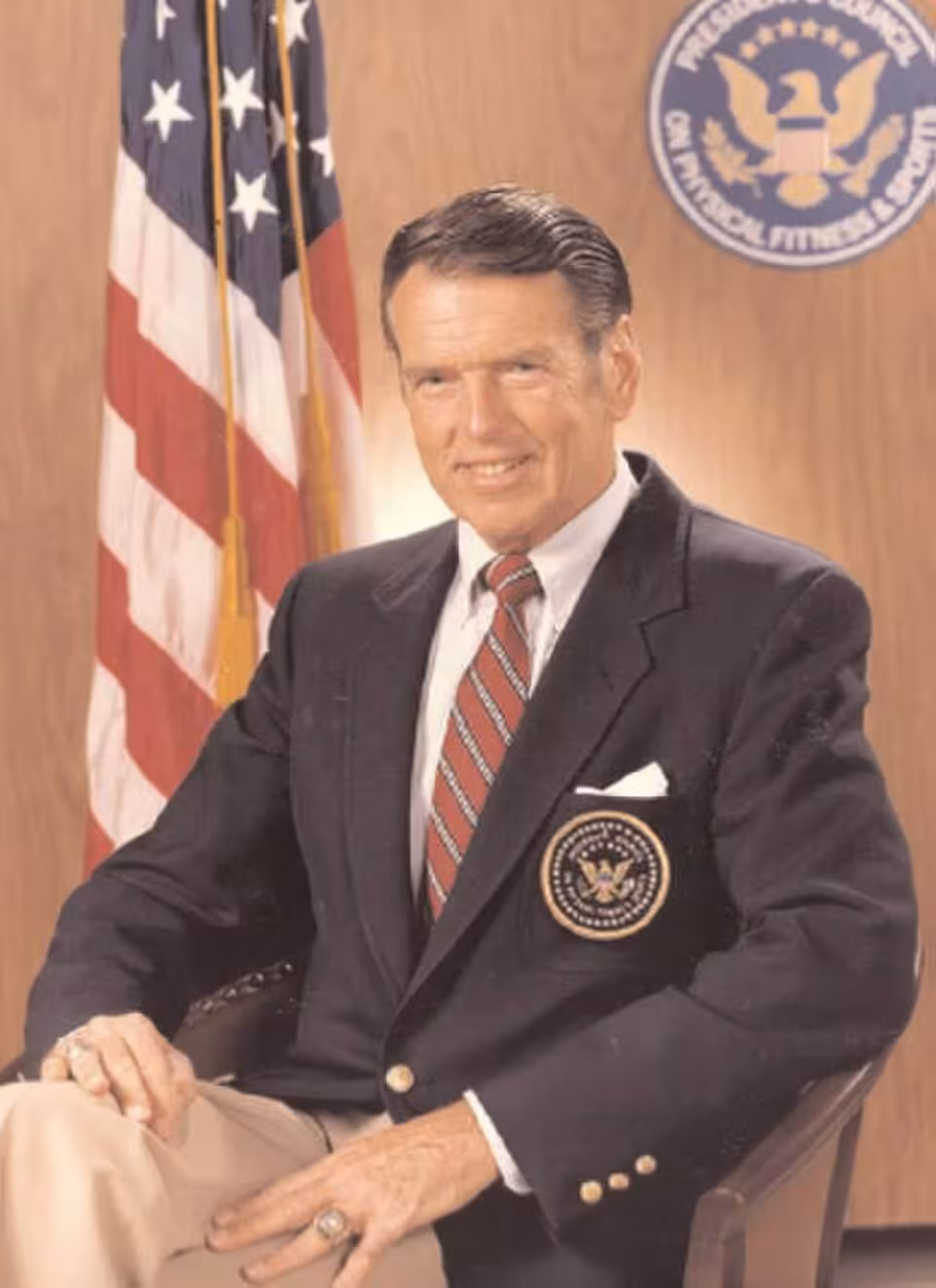 Color publicity photo of George Allen posing in a black sports coat in front of an American flag.