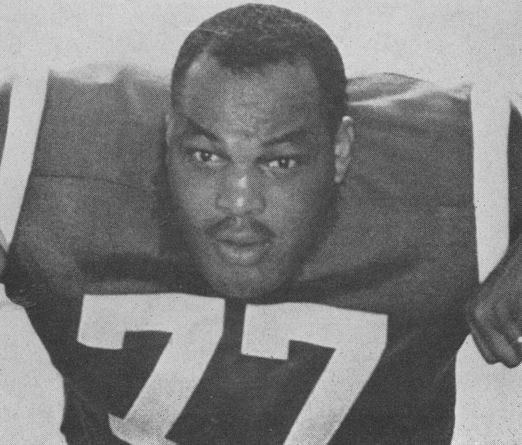 A black-and-white archival headshot of Baltimore Colts offensive lineman Jim Parker from 1960. The photo features a close-up of Parker in a three-point stance or leaning forward, wearing his jersey with the large white number '77' clearly visible. He has a focused, intense expression. The image has a grainy texture, characteristic of vintage newspaper or sports program halftone printing.