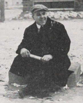 Archival candid of Knute Rockne in a winter overcoat and flat cap, holding a rolled document on the Notre Dame campus.