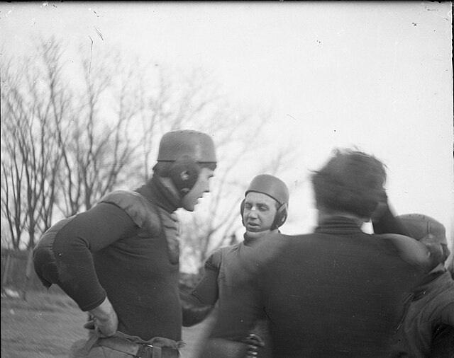 The black-and-white grainy image shows Heffelfinger in a group setting, likely during his time coaching or playing in an exhibition later in his career. He is the prominent figure on the left, standing with his hands on his hips. He wears a dark, long-sleeved jersey with reinforced padding on the shoulders and elbows. His headgear is a primitive, hard-shell leather helmet with ear flaps. Another player faces him, showing a bandage on his nose—a common sight in the "Iron Man" era.