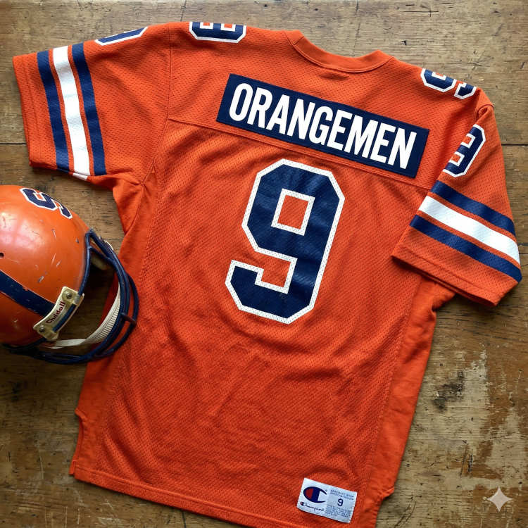 An orange Syracuse football jersey featuring the number '9' in blue with a white outline. The nameplate on the back reads 'ORANGEMEN' in white block letters on a blue background. The jersey is laid flat on a rustic wooden table next to a scuffed orange Riddell helmet with a blue face mask. A vintage Champion jock tag is visible at the bottom hem.