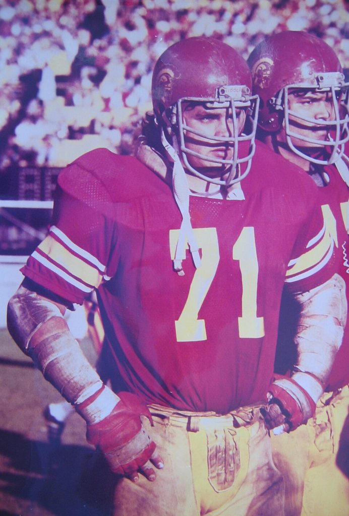 Color late 1970s image of Brad Budde palying for USC Football in between plays wearing his crimsom USC home jersey number 71.