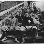 Vintage 1895–1896 sepia-toned black-and-white photograph of an early American football team sitting on the brick steps of South School. Features players wearing heavy quilted trousers, high-top lace-up leather boots, and dark turtlenecks with a stylized white emblem, including an African American player on the front left. A primary source for late-19th-century gridiron history from the MOHAI collection.