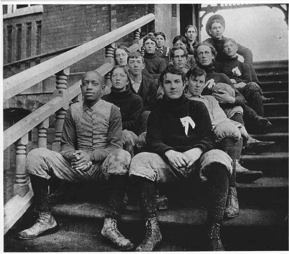 Vintage 1895–1896 sepia-toned black-and-white photograph of an early American football team sitting on the brick steps of South School. Features players wearing heavy quilted trousers, high-top lace-up leather boots, and dark turtlenecks with a stylized white emblem, including an African American player on the front left. A primary source for late-19th-century gridiron history from the MOHAI collection.
