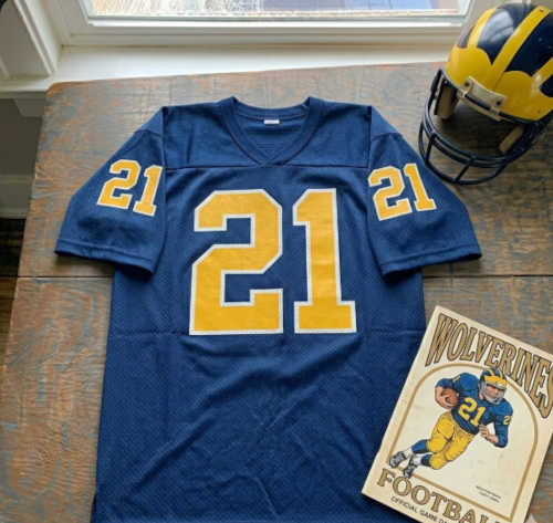 Vintage Michigan Wolverines #21 football jersey from Screenshot from 2026-05-12 04-46-41.jpg. Navy blue mesh with maize and white bordered block numbers. Includes the iconic winged helmet and a vintage Wolverines football program for historical Big Ten research