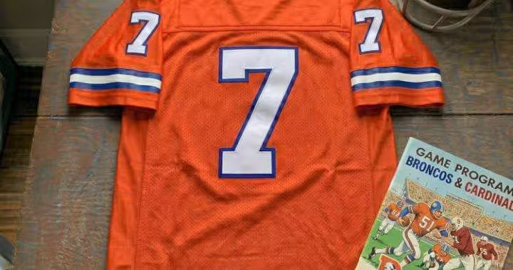 Vintage Denver Broncos #7 football jersey. Orange mesh with white and royal blue shadowed block numbers and striped sleeves. Features a Denver Broncos vs. Stanford Cardinal game program and a vintage leather helmet for historical NFL and John Elway research for Pigskin Dispatch archives.