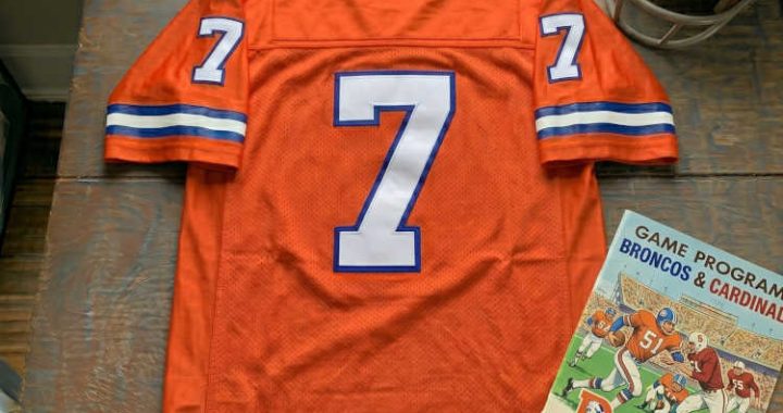 Vintage Denver Broncos #7 football jersey. Orange mesh with white and royal blue shadowed block numbers and striped sleeves. Features a Denver Broncos vs. Stanford Cardinal game program and a vintage leather helmet for historical NFL and John Elway research for Pigskin Dispatch archives.