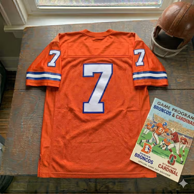 Vintage Denver Broncos #7 football jersey. Orange mesh with white and royal blue shadowed block numbers and striped sleeves. Features a Denver Broncos vs. Stanford Cardinal game program and a vintage leather helmet for historical NFL and John Elway research for Pigskin Dispatch archives.