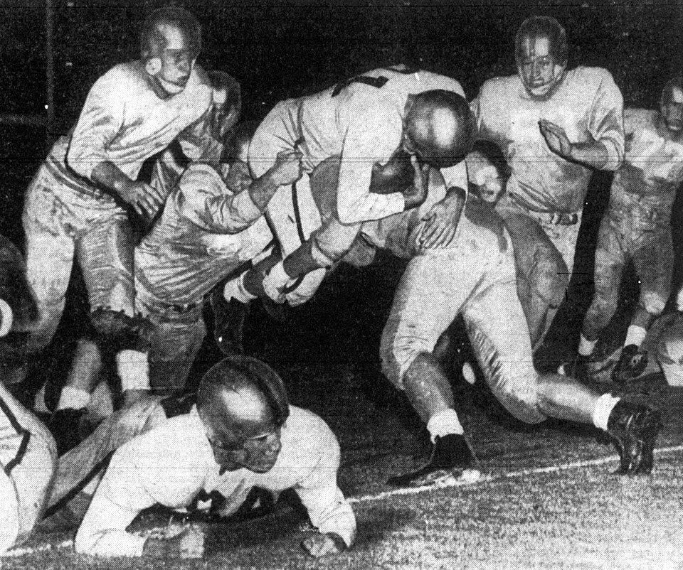black and white newspaper game action photo of Kansas Jayhawks halfback Don Hess (center, with ball) attempts to leap over the tackling UCLA Bruins defender Bob Heydenfeldt during a game on September 25, 1953 at the Los Angeles Memorial Coliseum in Los Angeles, California.