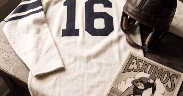 Black and white image of a white number 16 football jersey with a leather helmet and a 1926 Duluth Eskimos program sitting on a wooden table.