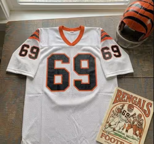 Vintage Cincinnati Bengals #69 football jersey as seen in Krumrie_69.jpg. White mesh with black and orange shadowed block numbers and tiger-striped sleeves. Includes a striped Bengals helmet and a vintage gameday program for historical NFL research on nose tackle Tim Krumrie for Pigskin Dispatch archives.