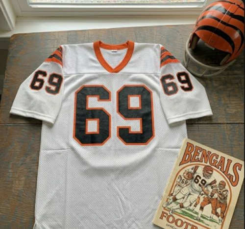 Vintage Cincinnati Bengals #69 football jersey as seen in Krumrie_69.jpg. White mesh with black and orange shadowed block numbers and tiger-striped sleeves. Includes a striped Bengals helmet and a vintage gameday program for historical NFL research on nose tackle Tim Krumrie for Pigskin Dispatch archives.