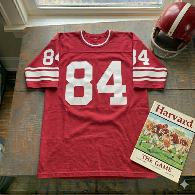 Vintage Harvard Crimson #84 football jersey. Crimson fabric with large white block numbers and double-striped sleeves. Features a Harvard 'The Game' official program and a crimson helmet with a white stripe for Ivy League history and Pigskin Dispatch archives.