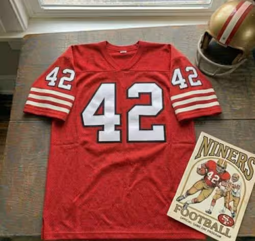 Vintage San Francisco 49ers #42 football jersey. Scarlet mesh with white and black shadowed block numbers and triple-striped sleeves. Features a 49ers gameday program and a gold helmet with red stripes for historical NFL research on Hall of Fame safety Ronnie Lott