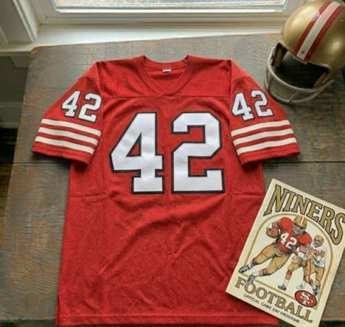 Vintage San Francisco 49ers #42 football jersey. Scarlet mesh with white and black shadowed block numbers and triple-striped sleeves. Features a 49ers gameday program and a gold helmet with red stripes for historical NFL research on Hall of Fame safety Ronnie Lott