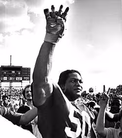 Black and white image of Simmons while playing for Florida State University in 1981 raising his right hand with four fingers to signat the start of the fourth quarter during a 1981 FSU game.
