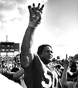 Black and white image of Simmons while playing for Florida State University in 1981 raising his right hand with four fingers to signat the start of the fourth quarter during a 1981 FSU game.