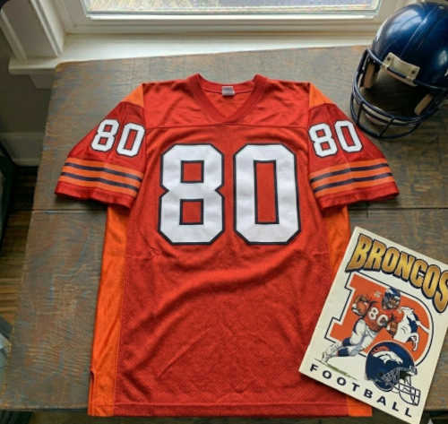 Vintage Denver Broncos #80 football jersey as seen in SMith_80.jpg. Orange mesh with white and navy blue shadowed block numbers. Includes a navy blue Broncos helmet and a vintage Broncos football program for historical NFL research and Pigskin Dispatch archives.