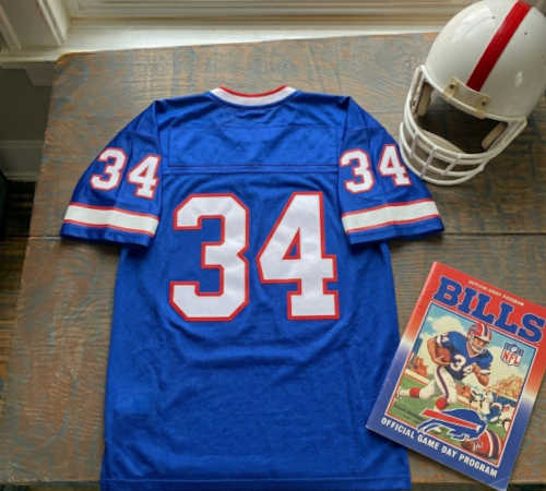 A 1990s era Blue Buffalo Bills style number 34 jersey displayed on a wooden table with a white helmet , with red stripe, and a vintage Bill Program.