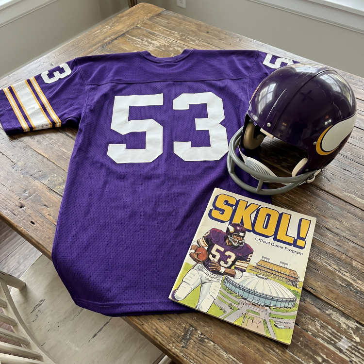 Color image of a 1970s era Minnesota NFL football jersey 53, without a name on it, and matching helmet. The program cover should say Skol!