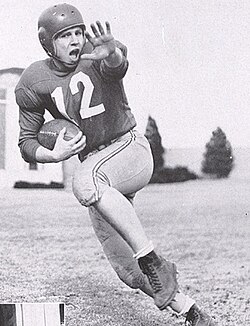 Vintage 1950 black-and-white action photograph from Bobby_Reynolds_(American_football).jpg featuring Nebraska Cornhuskers All-American halfback Bobby Reynolds (#12) executing a stiff arm during a run. Reynolds wears a dark jersey with white block numbers, light-colored pants with a side stripe, high-top cleated leather shoes, and a molded plastic helmet without a facemask. A primary source for Big Seven college football history and Pigskin Dispatch archives."