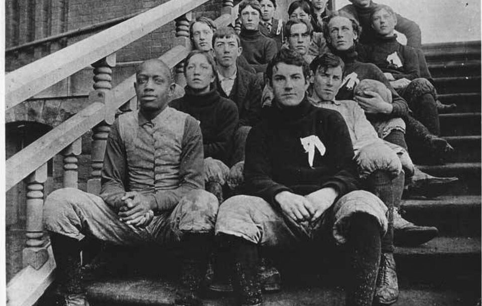 Vintage black and white image of a late nuneteenth century American football high school integrated team posing for a photo on some prominent cement steps.