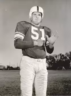 Vintage 1954 black-and-white promotional photograph from Kurt_Burris.png featuring Oklahoma Sooners All-American center Kurt Burris (#51) posing on a practice field. He wears a dark jersey with white block numbers and double sleeve stripes, light-colored pants, and a white molded plastic helmet with a dark center stripe and no facemask. A primary source for Big Seven college football history and Pigskin Dispatch archives.