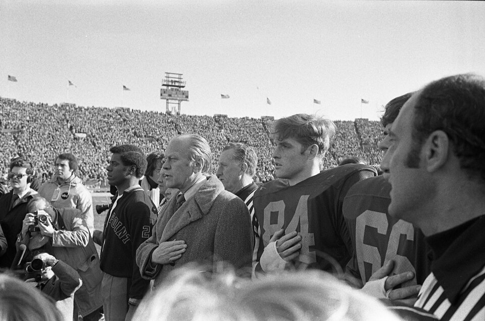 Black and white image of This photograph depicts President Gerald R. Ford and others standing with their hands over their hearts for the National Anthem prior to the start of the 75th Army-Navy football game at John F. Kennedy Stadium in Philadelphia, Pennsylvania. Pictured from left are Army team captain Robert Johnson, President Ford, and Navy team captains Cliff Collier and Tim Harden. Unknown author or not provided