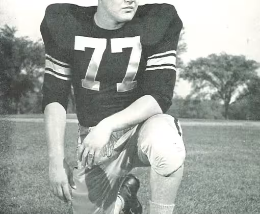 Full body publicity yearbook pose of Alaen Karras in Iowa uniform kneeling with helmet off wearing number 77 in 1958.