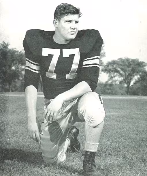 Full body publicity yearbook pose of Alaen Karras in Iowa uniform kneeling with helmet off wearing number 77 in 1958.