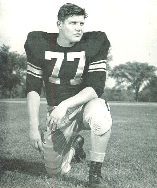 Full body publicity yearbook pose of Alaen Karras in Iowa uniform kneeling with helmet off wearing number 77 in 1958.