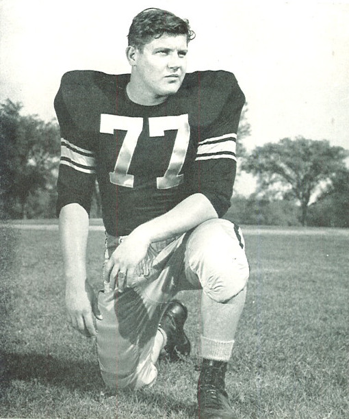 Full body publicity yearbook pose of Alaen Karras in Iowa uniform kneeling with helmet off wearing number 77 in 1958.
