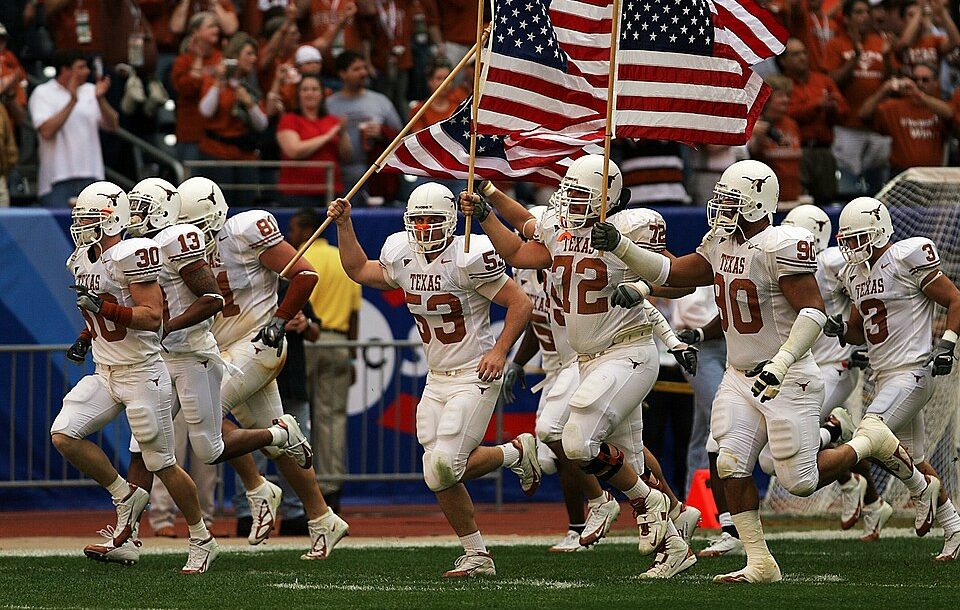 Full color photo of white clad University of Texas Longhorns football players enter the field for the 2005 Bix XI Championship Game., one carrying an American flag KeithJJ on Pixabay