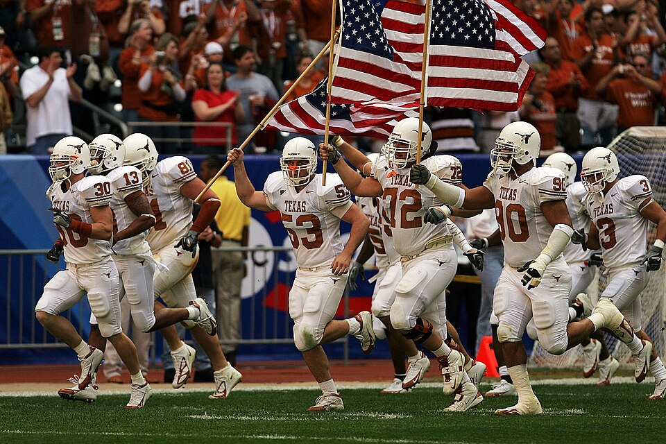 Full color photo of white clad University of Texas Longhorns football players enter the field for the 2005 Bix XI Championship Game., one carrying an American flag KeithJJ on Pixabay