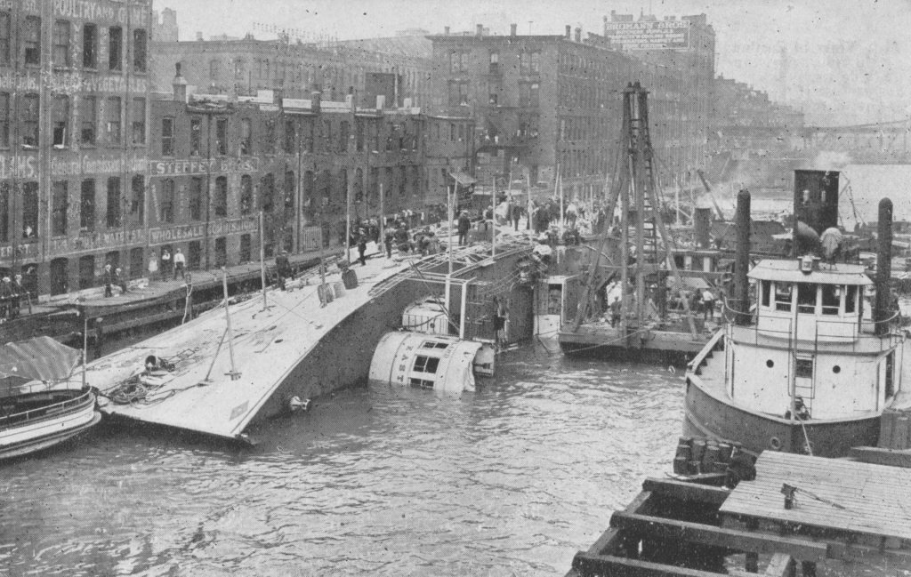 Black and white vintage photo of the Eastland taken from Fire Tug in river, showing the hull resting on its side on the river bottom. The work of recovering bodies is being carried on with untiring effort.